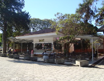 Cafeteria com mesas externas e toldos, cercada por árvores, em um dia ensolarado.
