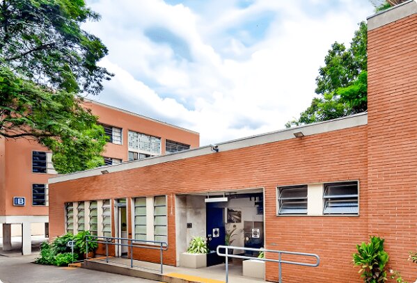 Edifício de tijolos com janelas em brise e rampa de acesso, em frente a um prédio laranja com janelas.