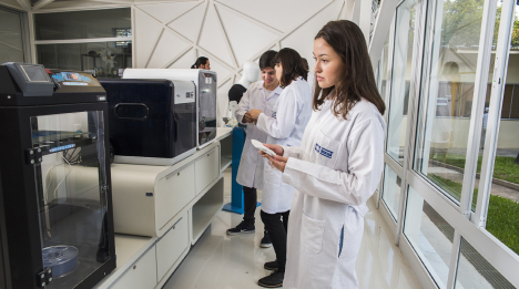 Estudantes em jaleco branco observam impressoras 3D em laboratório do Instituto Mauá de Tecnologia.