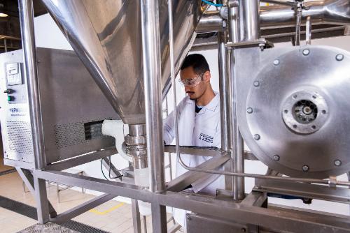 Homem de jaleco e óculos de segurança opera máquina industrial em laboratório do Instituto Mauá de Tecnologia.