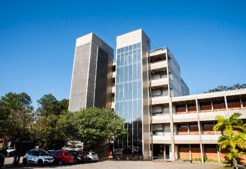 Edifício moderno com fachada de vidro e concreto, cercado por árvores e com carros estacionados.