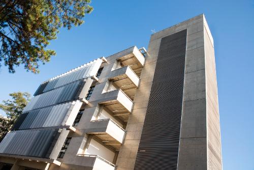 Edifício moderno de concreto com varandas e painéis metálicos sob céu azul.