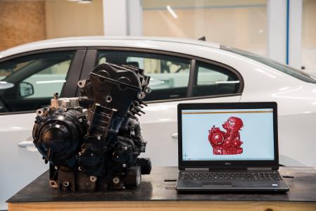 Motor de motocicleta preto ao lado de um laptop exibindo um modelo 3D vermelho.