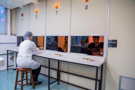 Pessoas em cabines separadas, uma com jaleco e touca, outra com um homem e uma mulher visíveis através de janelas.