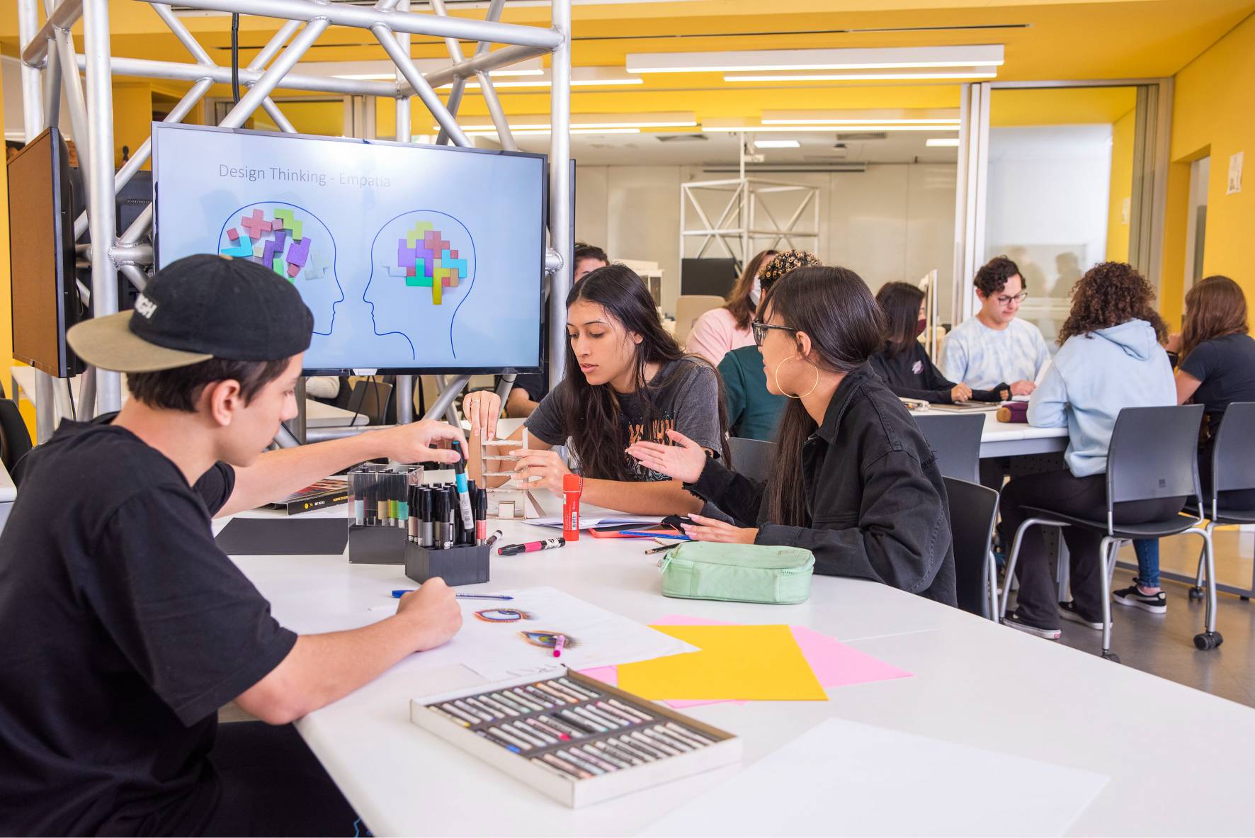 Estudantes em sala de aula do Instituto Mauá de Tecnologia trabalham em projetos, com tela exibindo "Design Thinking - Empatia".