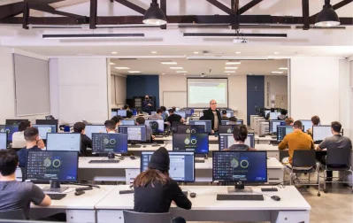 Sala de aula com alunos e professor em frente a computadores, com projeção de tela ao fundo.
