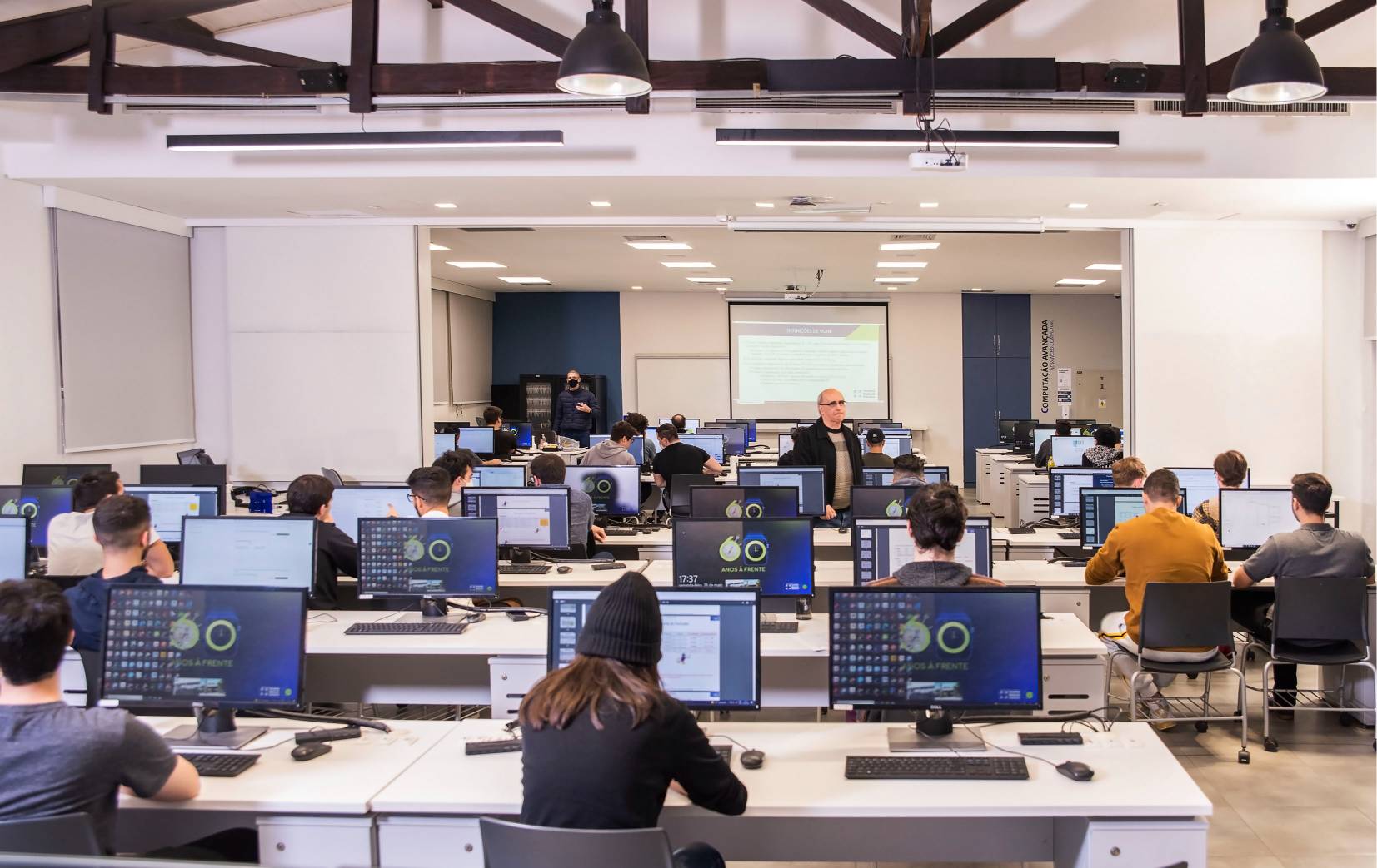 Sala de aula com alunos e professor em frente a computadores, com projeção de tela ao fundo.