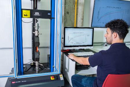 Homem opera máquina de ensaio de materiais com gráficos em monitores.