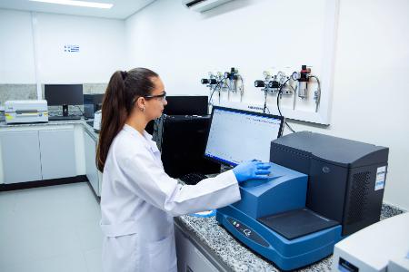 Mulher de jaleco e luvas azuis opera equipamento em laboratório.