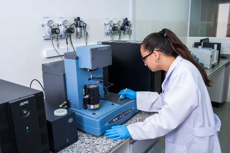 Mulher de jaleco e luvas azuis opera equipamento de laboratório azul, com painel de controle e amostra em foco.