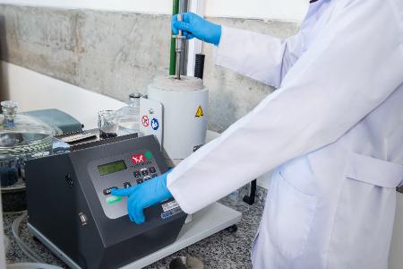 Pessoa de jaleco branco e luvas azuis opera equipamento de laboratório com tela digital.