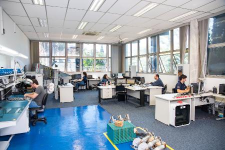 Laboratório com pessoas trabalhando em bancadas com equipamentos eletrônicos e computadores.