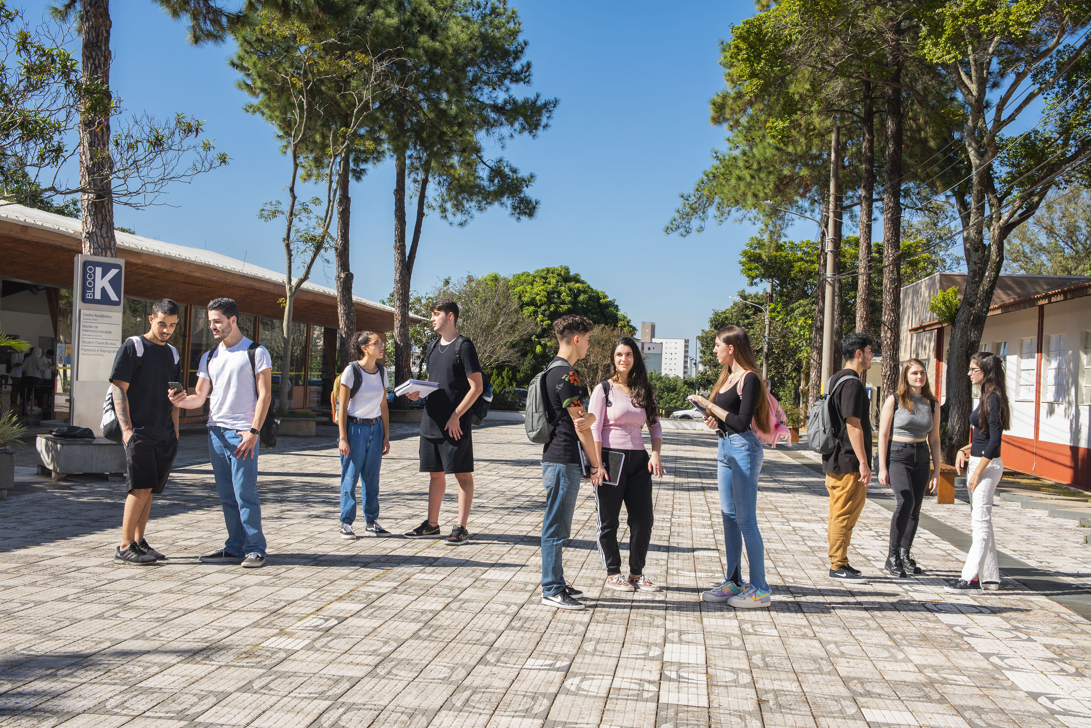 Estudantes do Instituto Mauá de Tecnologia conversam em um pátio ensolarado com prédios e árvores ao fundo.