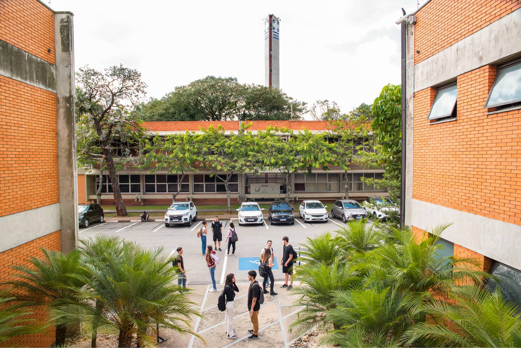 Estudantes em um estacionamento no Instituto Mauá de Tecnologia, com carros e um prédio ao fundo.