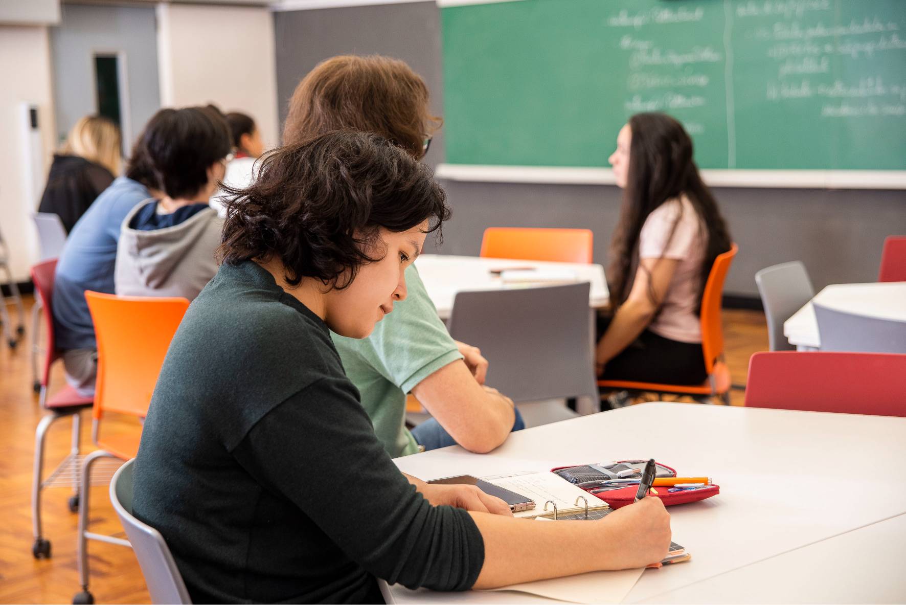 Estudantes em sala de aula, um escrevendo em um caderno, com quadro verde ao fundo.