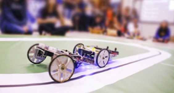 Dois robôs competem em uma pista, com espectadores desfocados ao fundo.