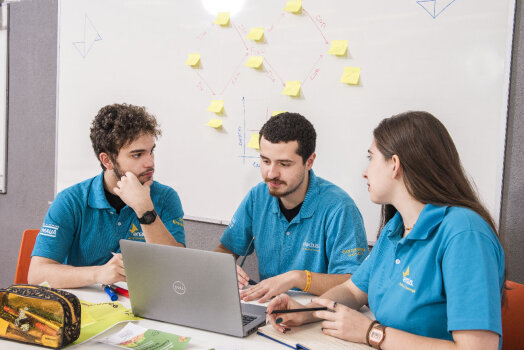 Três estudantes do Instituto Mauá de Tecnologia colaboram em um projeto com laptop e quadro branco.