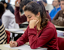 Estudante concentrada escrevendo em uma sala de aula, com outros alunos ao fundo.